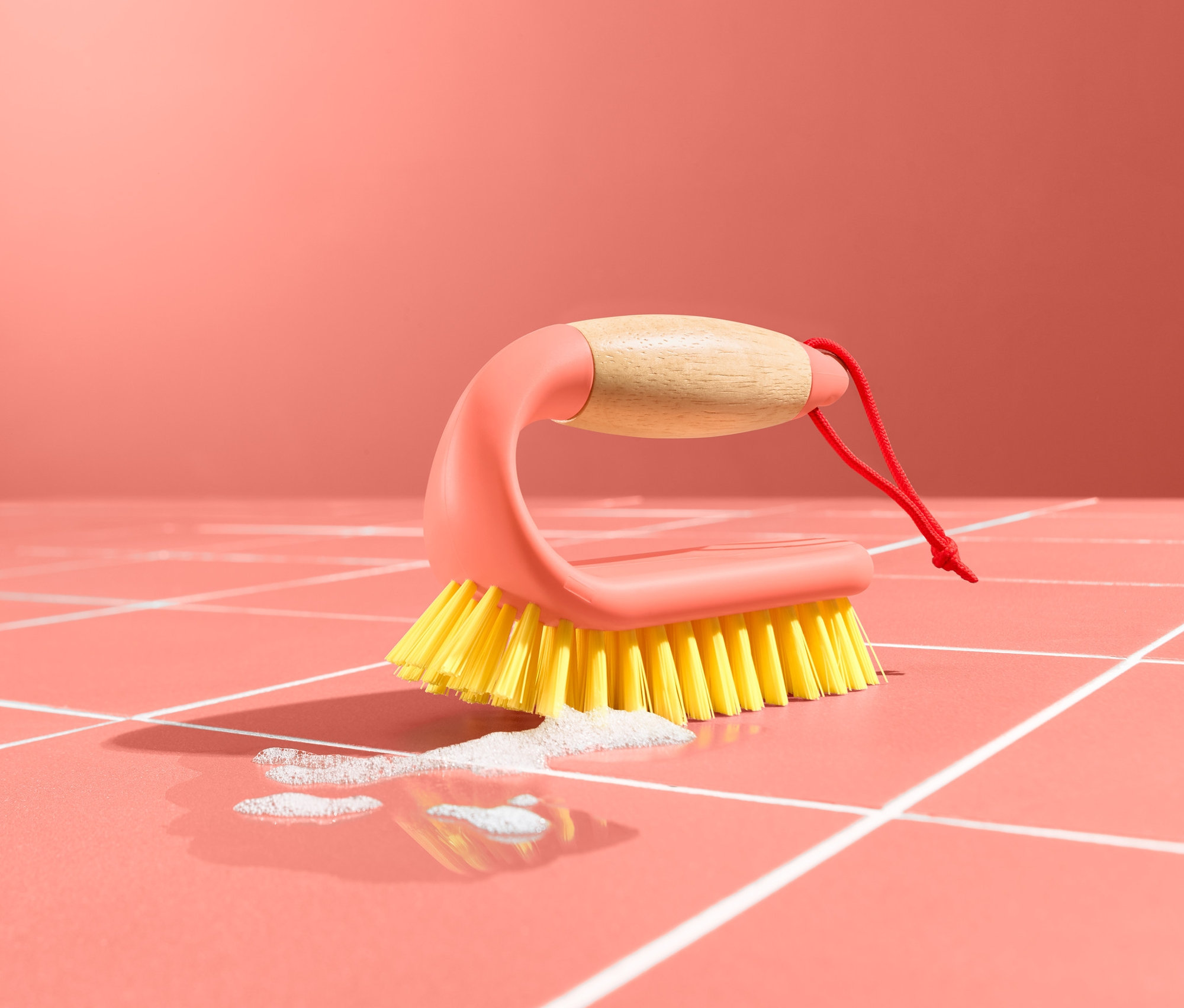 Egy csempe- és padlótisztító kefe fekszik egy rózsaszín, fehér fugás csempézett padlón, egy kis tócsával.