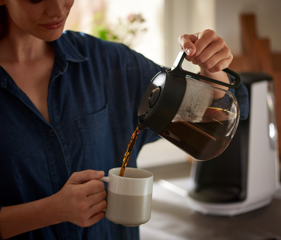 Nő kávét önt egy kancsóból egy bögrébe. A háttérben egy »Let's Brew« filteres kávéfőző látható.