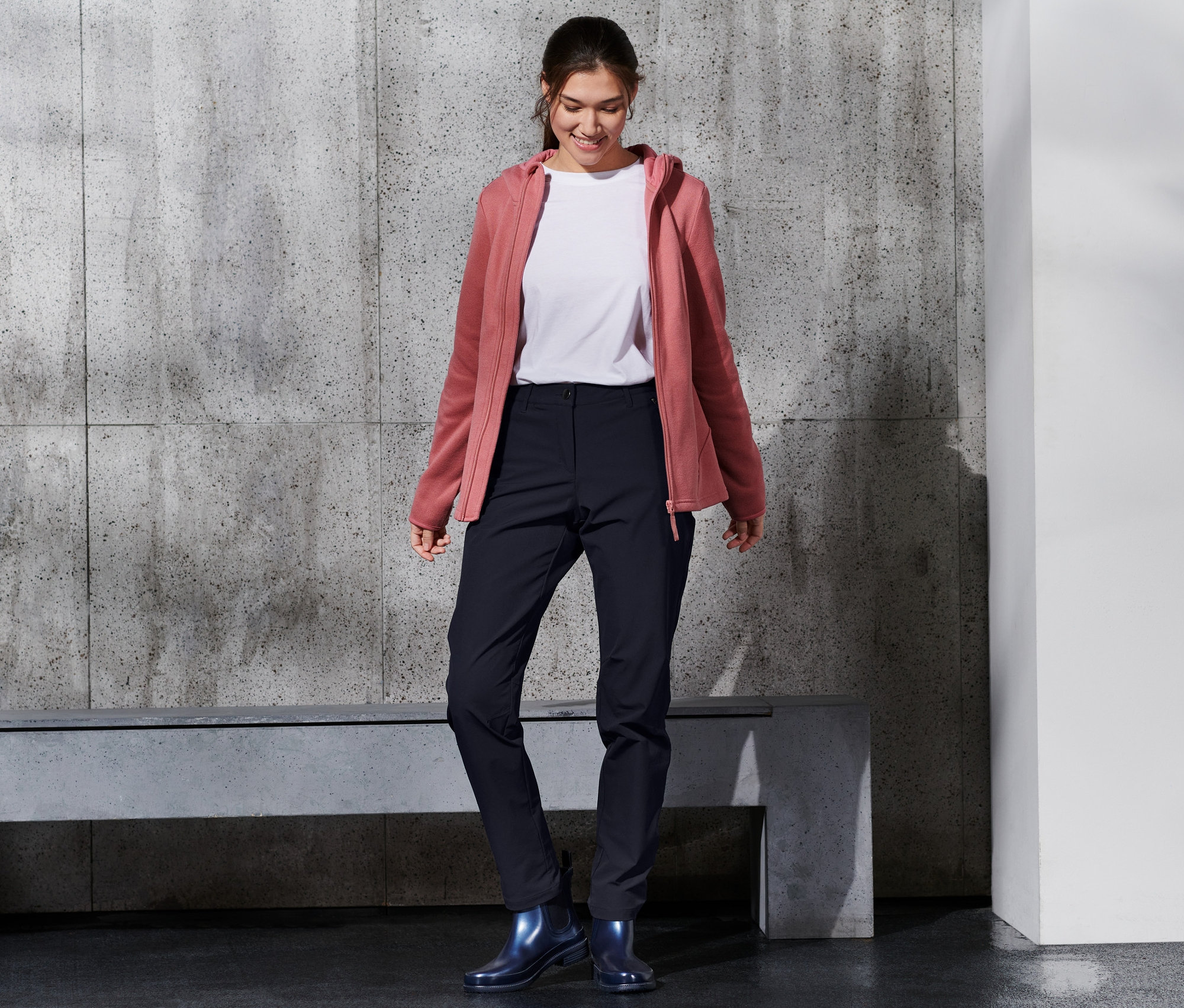 Mosolygó nő softshell nadrágot, fehér pólót és rózsaszín kapucnis pulóvert visel.