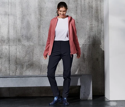 Mosolygó nő softshell nadrágot, fehér pólót és rózsaszín kapucnis pulóvert visel.