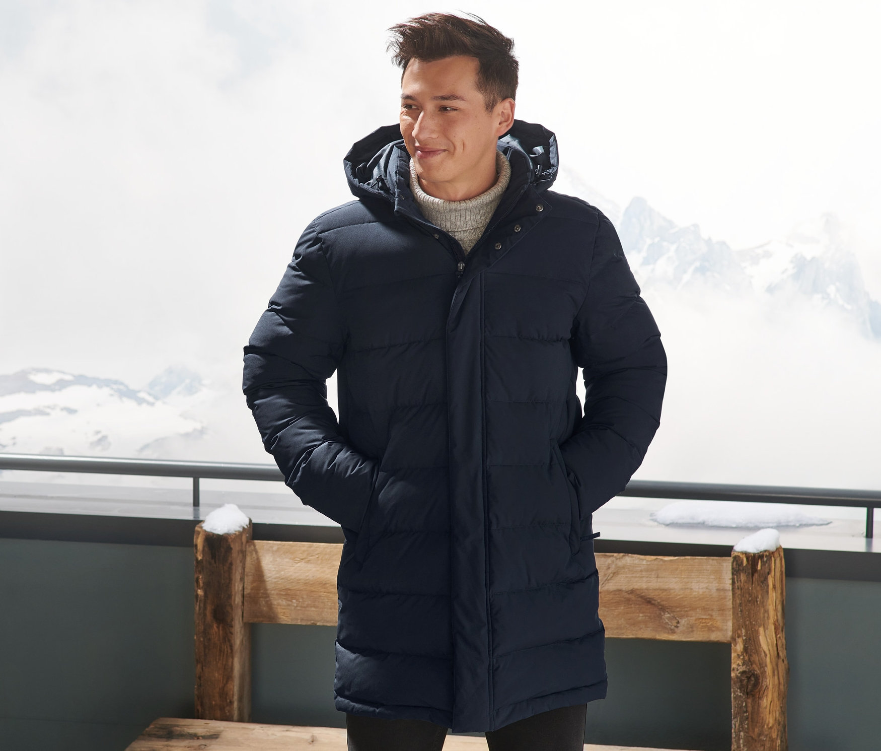 Egy férfi sötétkék színű, steppelt pehelykabátban áll kint, kezét a zsebébe dugva.