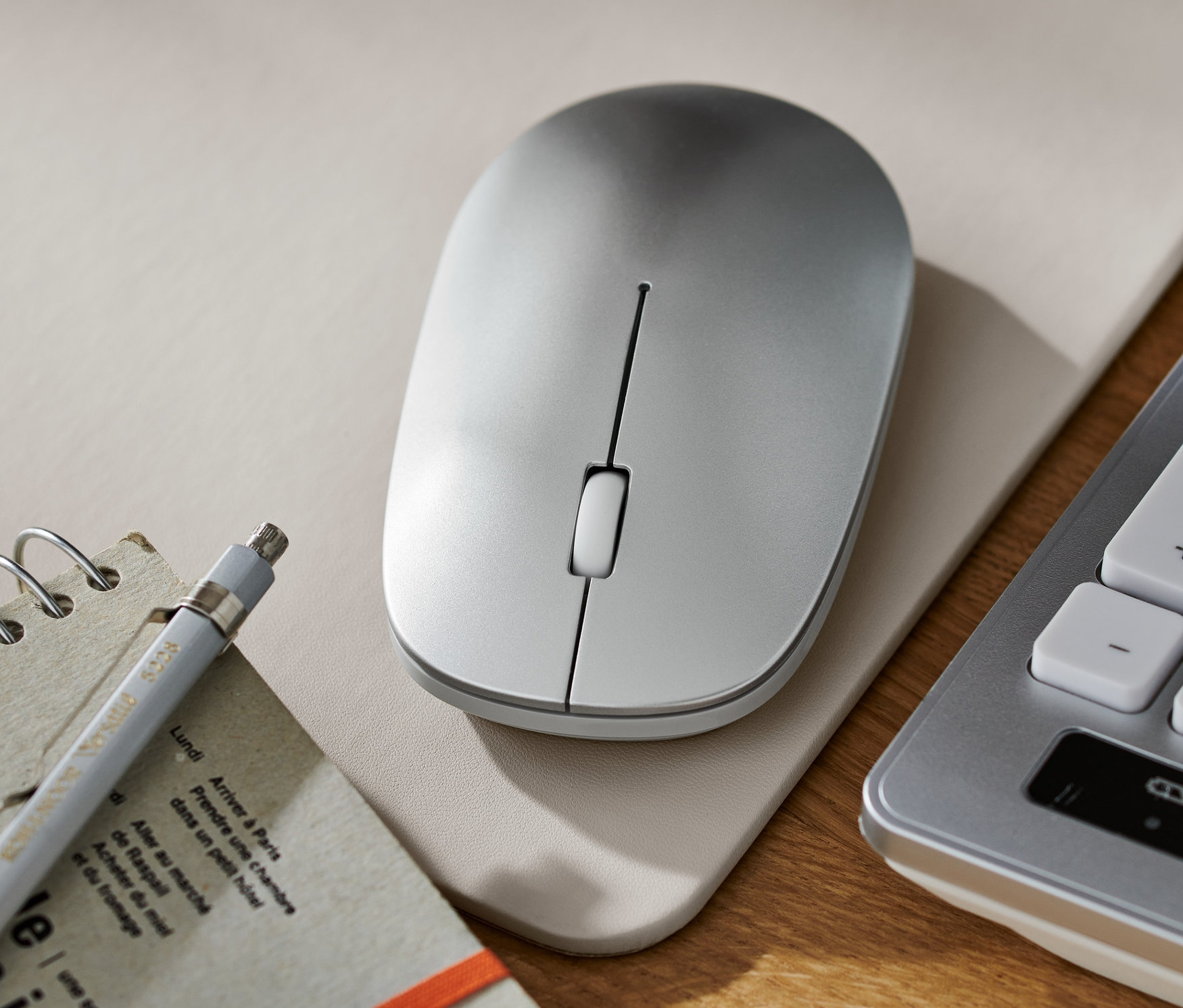 Egy ezüst vezeték nélküli egér fekszik egy alátéten egy jegyzettömb, egy ceruza és egy billentyűzet mellett.