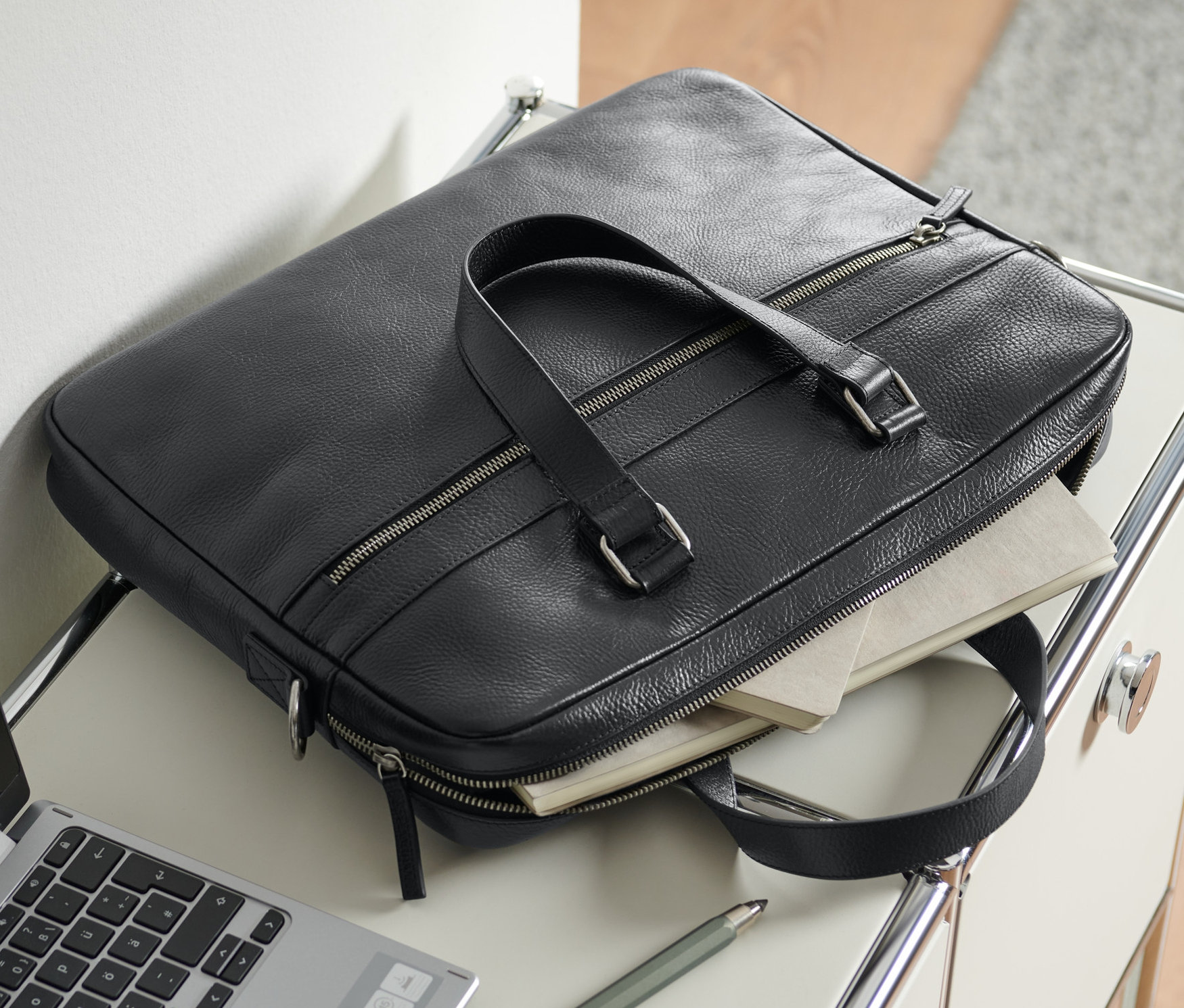 Egy fekete férfi bőr aktatáska egy asztalon fekszik egy laptop és egy toll mellett.