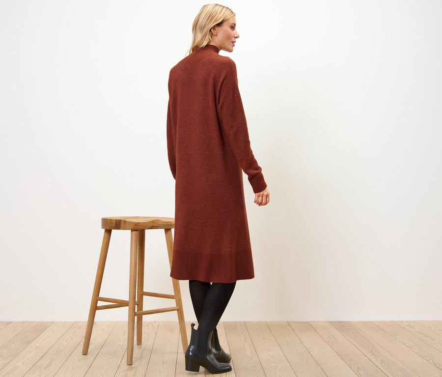 Egy nő oldalt áll, kötött, gyapjútartalmú ruhát, fekete harisnyát és bokacsizmát visel. Mellette egy fából készült szék áll.