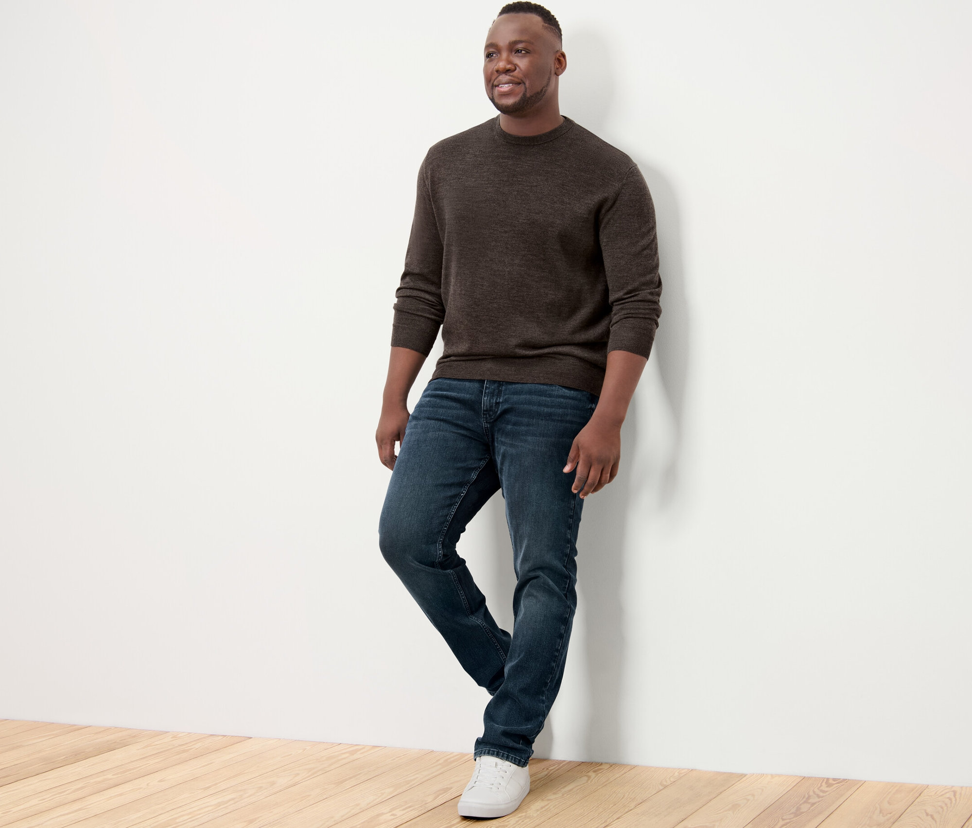 Férfi támaszkodik a falnak, barna merinó pulóvert, farmert és tornacipőt visel.