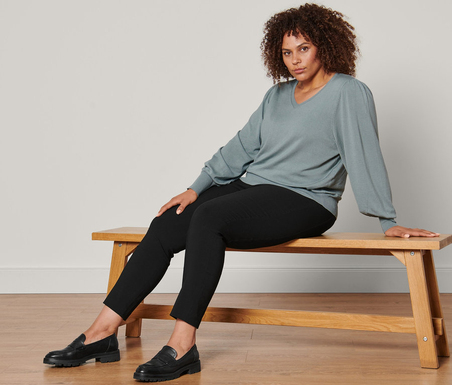 Nő ül egy padon, visel egy női finomkötésű pulóvert, sztreccsnadrágot és fekete bőr mokaszínt.