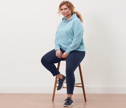 Nő ül egy széken, világoskék kapucnis szabadidőfelsőt, sötétkék sztreccsnadrágot és női sneakercipőt visel.