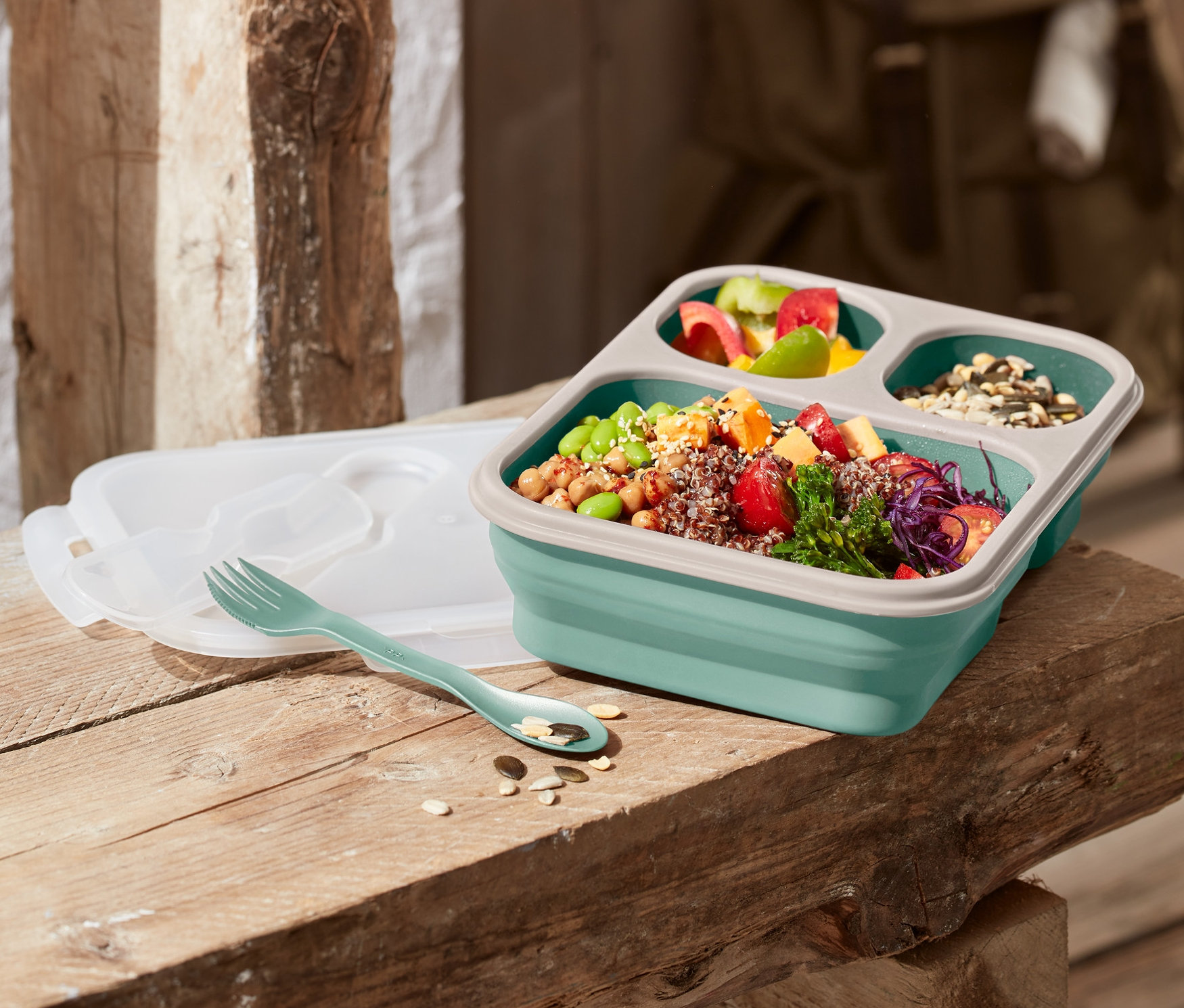 Egy összehajtható ételhordó doboz egy fa padon, mellette egy villa és egy fedő.