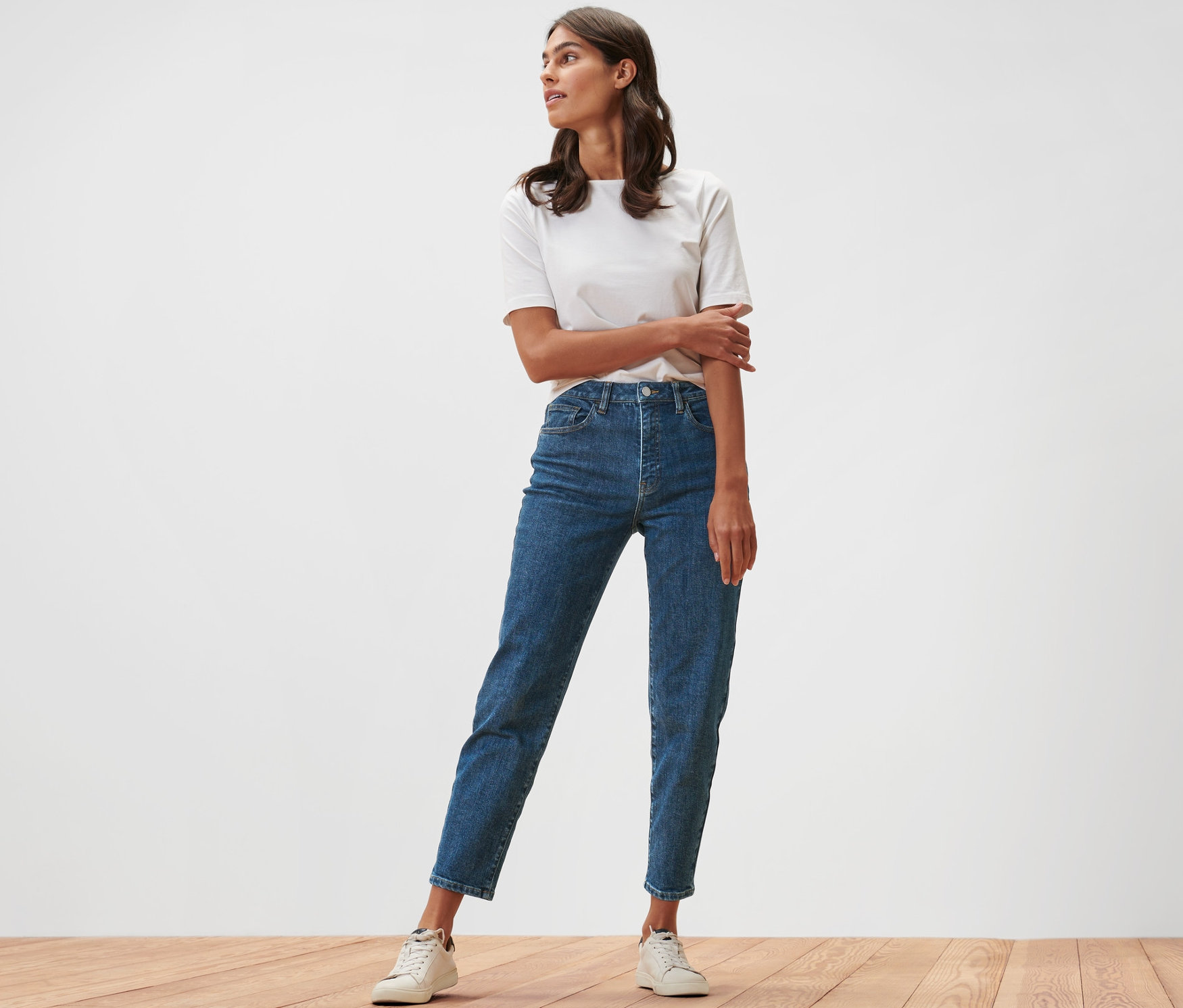 Egy nő áll egy fapadlón, Mom fit »Nyla« farmernadrágot és fehér pólót visel.