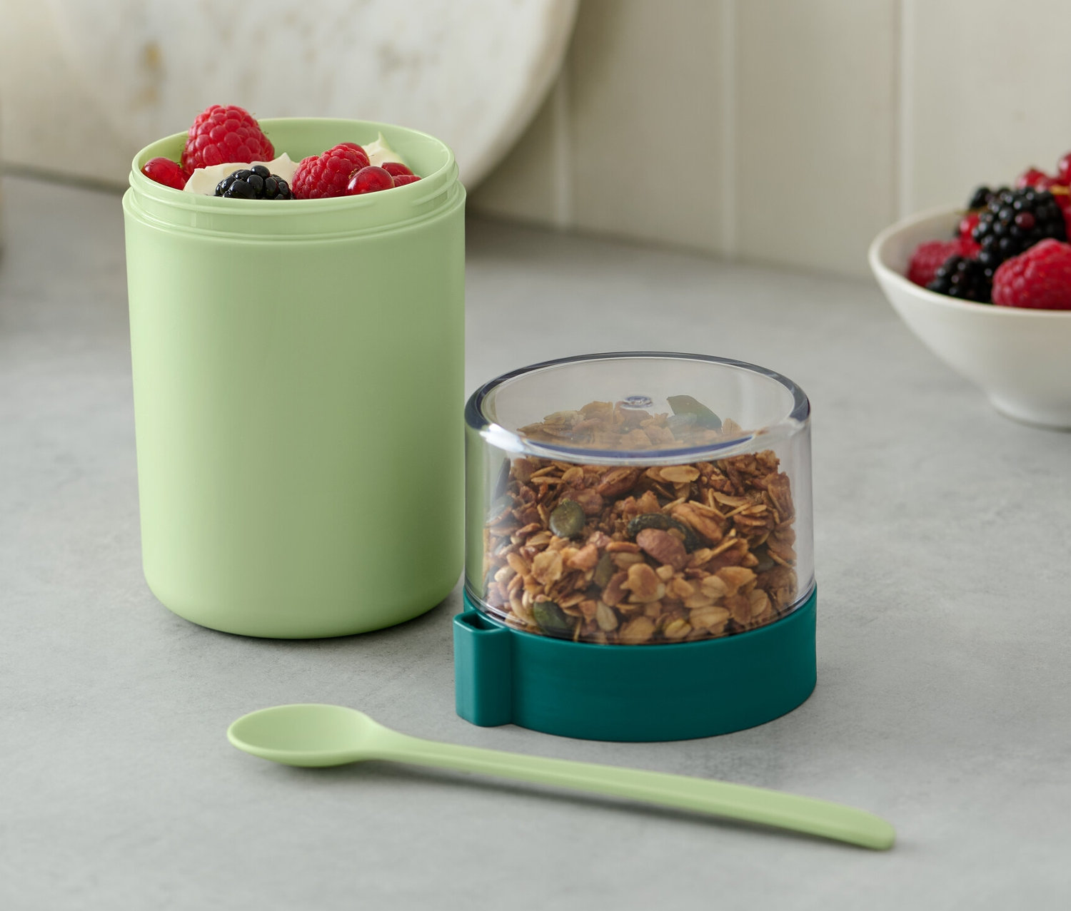 Egy zöld színű to-go joghurtos pohár müzlivel és bogyós gyümölcsökkel.