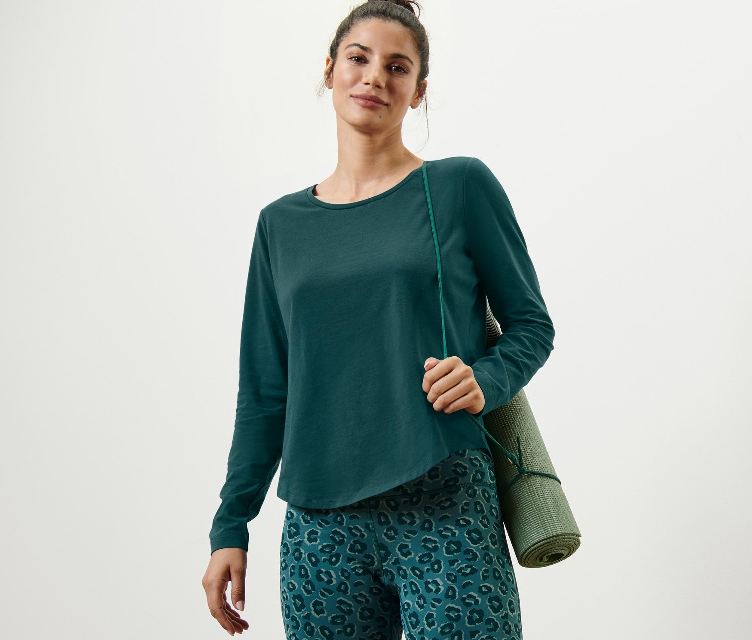 Nő visel egy zöld, hosszú ujjú sportfelsőt és egy zöld jógamatracot.