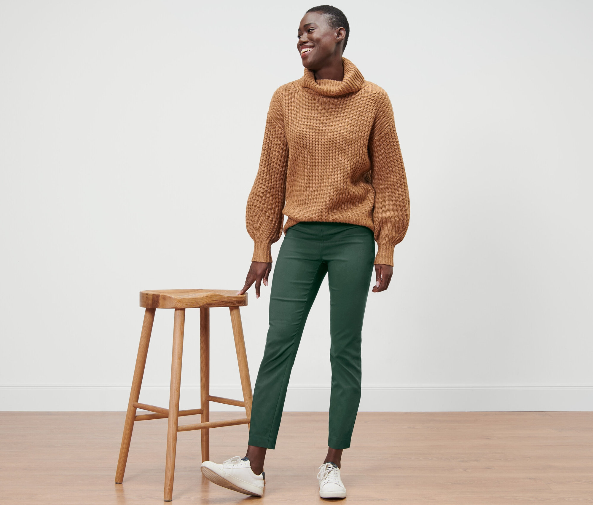 Egy mosolygó nő áll egy fa szék mellett, barna pulóvert és zöld sztreccsnadrágot visel.