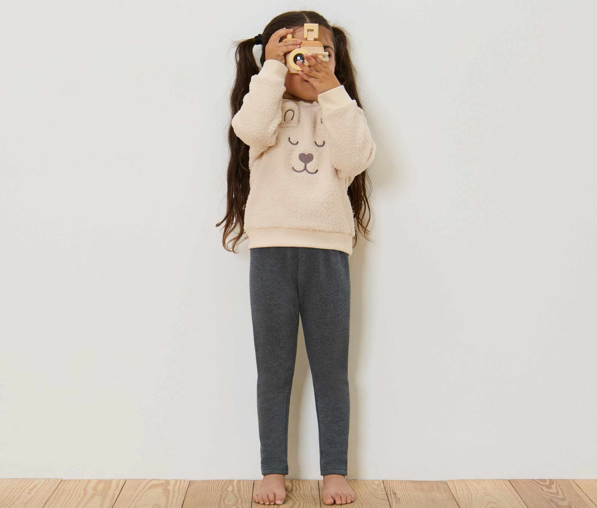 Kislány néz át egy fa fényképezőgépen, szürke gyerek termoleggingset visel.