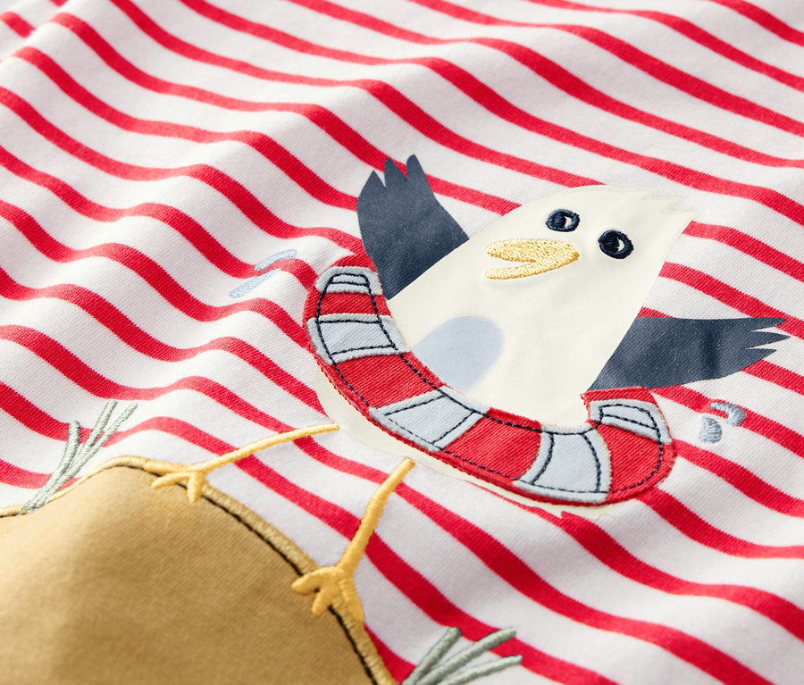 Egy piros csíkos, sirályos, úszógumis rátéttel ellátott gyerekpóló részlete.