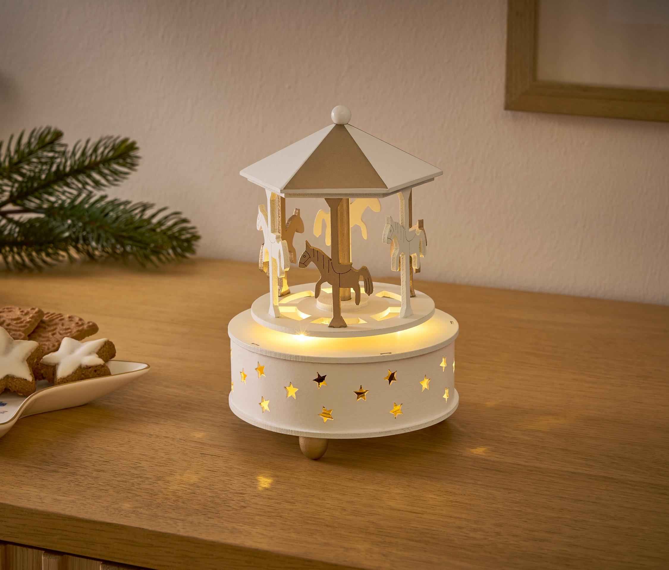 Egy fából készült körhinta lovakkal és csillagokkal világít egy fa asztalon karácsonyi kekszek mellett.