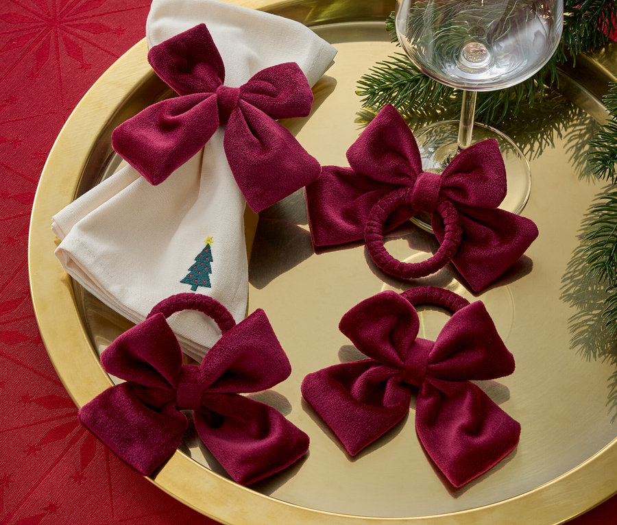 Egy arany tálcán karácsonyfa hímzéssel ellátott szövet szalvéták, bordó masnik és egy borospohár látható.