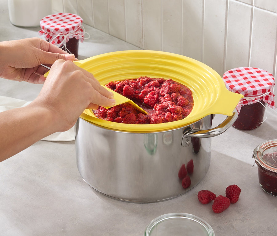 Egy személy málnadzsemet kever egy edényben sárga fröccsenésvédővel. Néhány málna és üveg áll a konyhapulton.