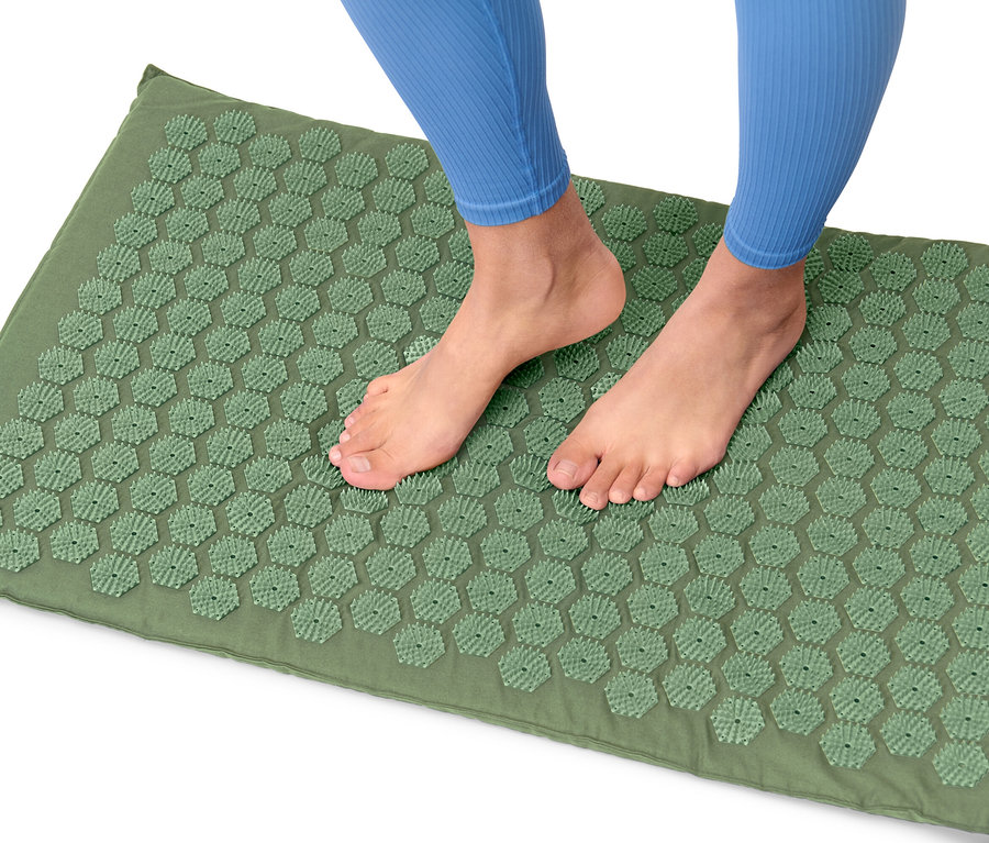 Egy kék leggingset viselő személy lábai egy zöld akupresszúrás matracon állnak.