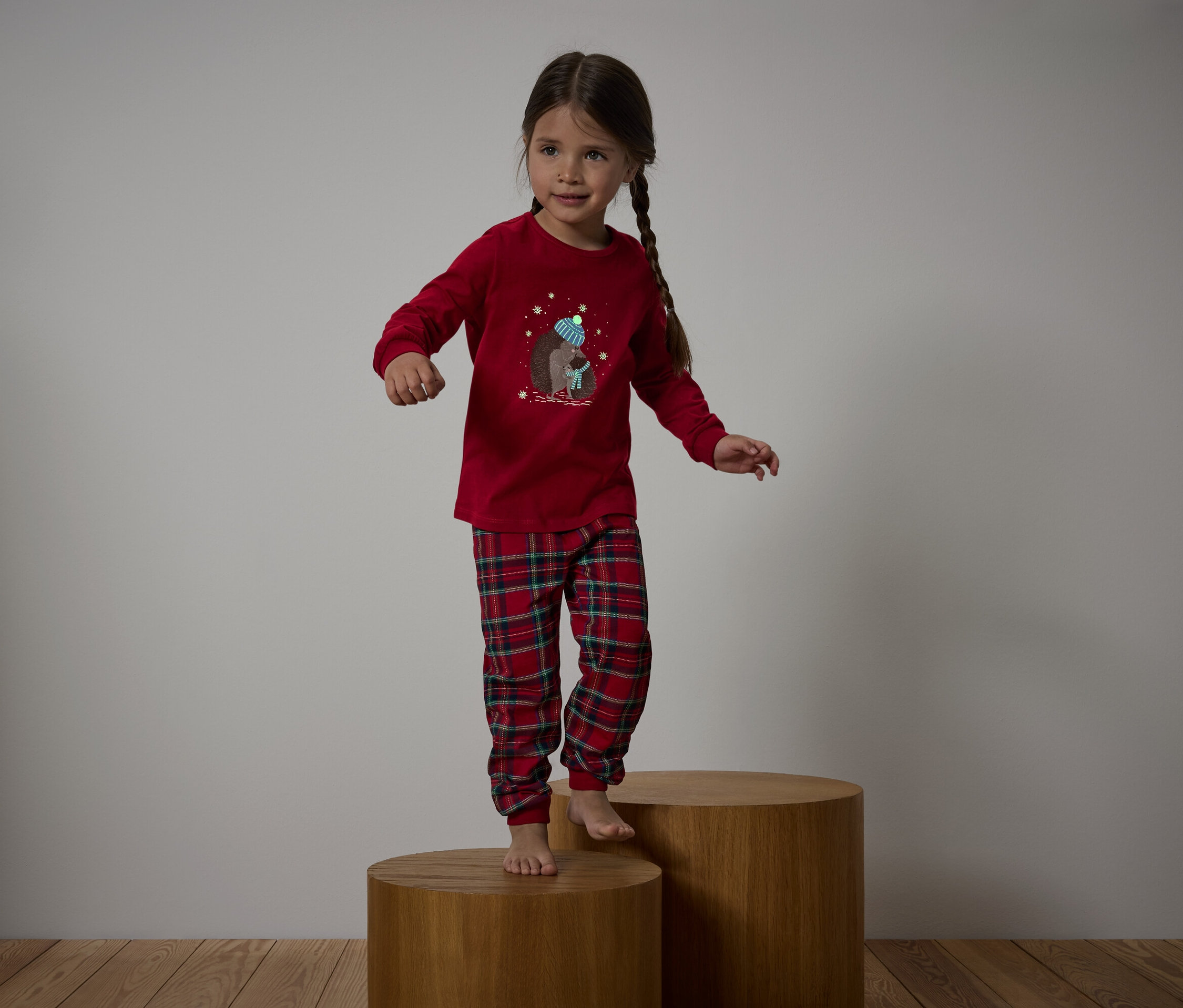 Egy lány piros pizsamában kockás nadrággal fa talapzaton áll.