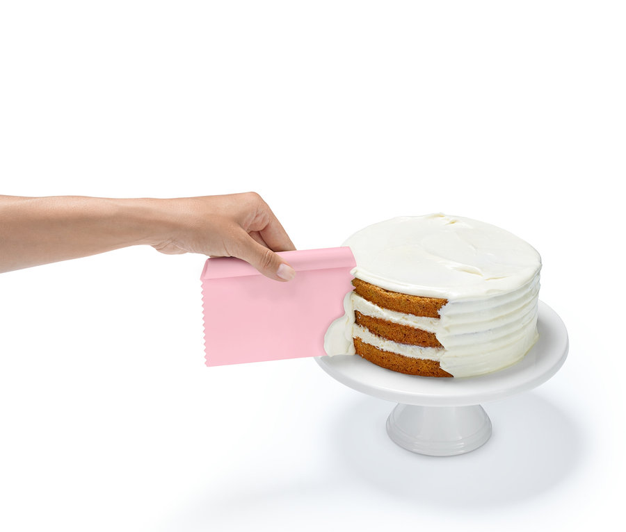 Egy kéz rózsaszín spatulával mázat ken egy háromrétegű tortára, ami egy fehér talapzaton áll.