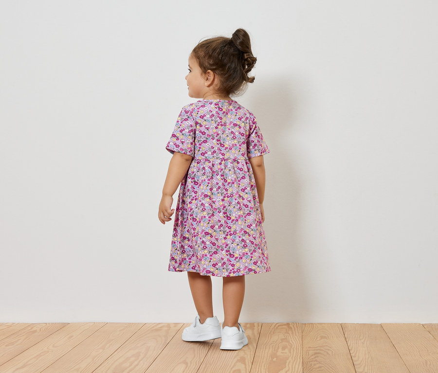 Virágmintás ruhát és fehér tornacipőt viselő kislány áll háttal a kamerának.