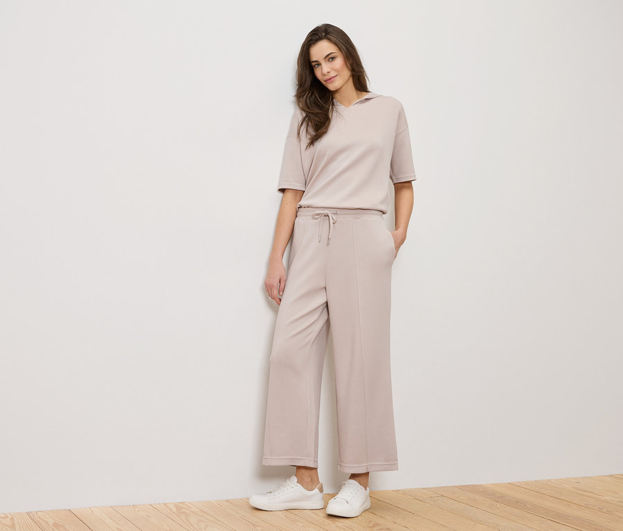 Egy nő áll egy fehér fal előtt, bézs színű ruhát és fehér tornacipőt visel.