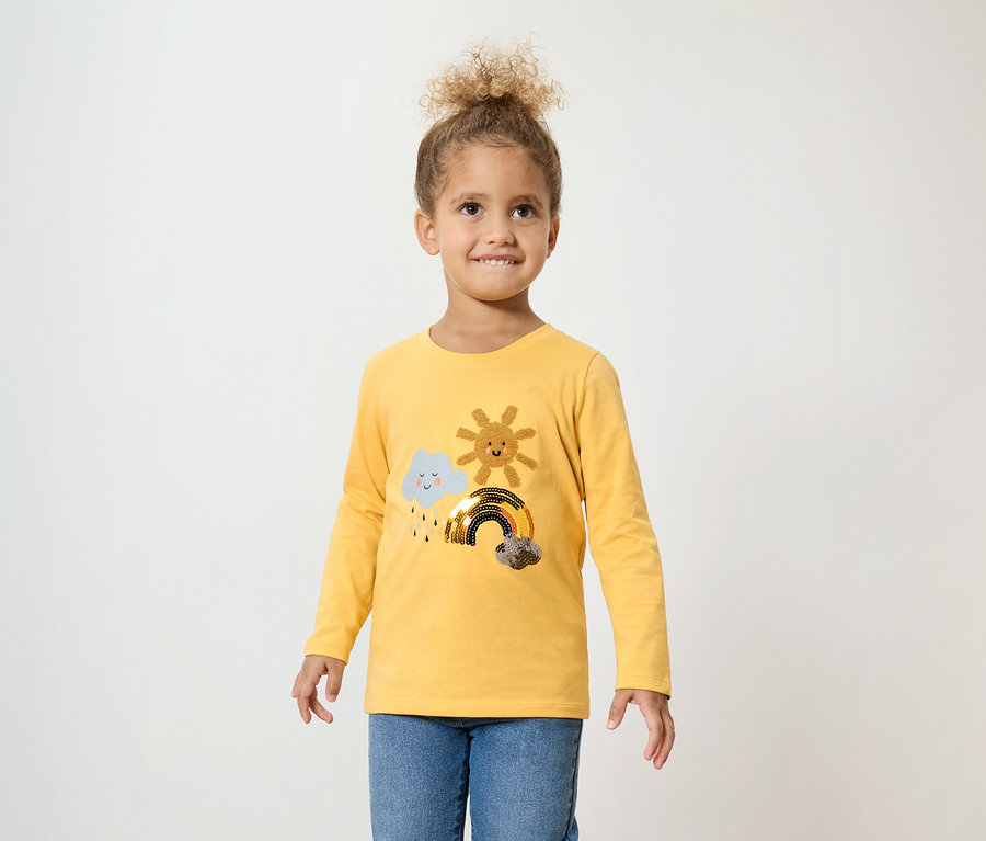 Göndör hajú lány felhővel, nappal és szivárvánnyal díszített sárga, hosszú ujjú pólót és kék farmert visel.