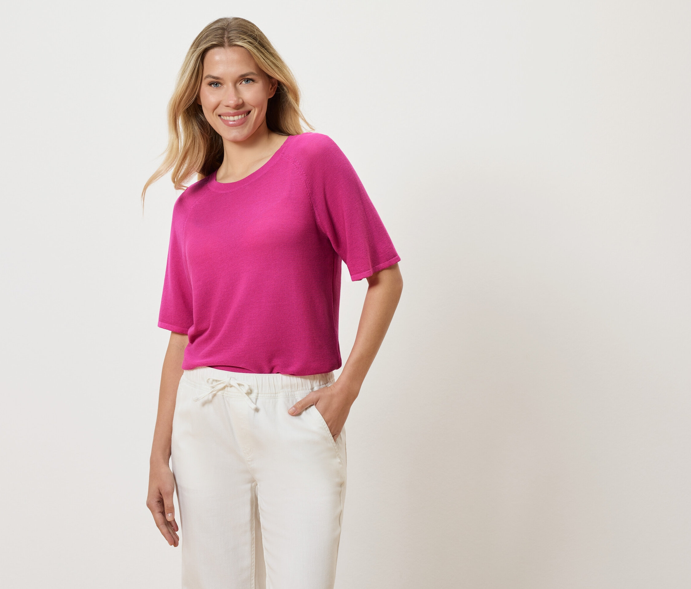 Egy szőke, hullámos hajú nő rózsaszín pólót és fehér nadrágot visel, és a kezét a zsebébe teszi.