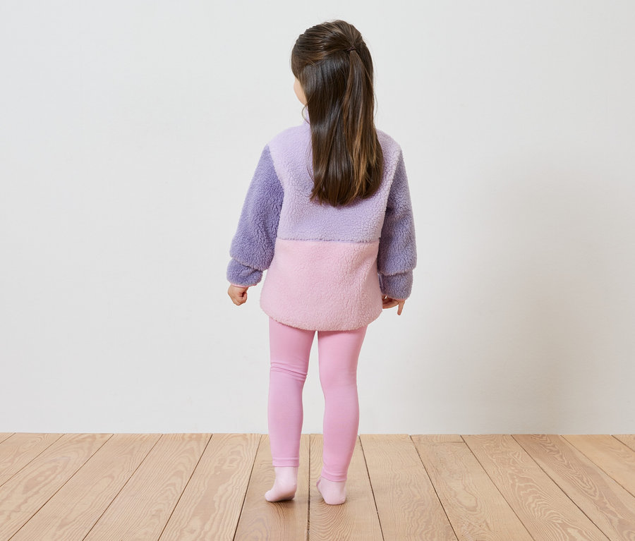 Hátulnézet egy hosszú, barna hajú lányról, aki levendula és rózsaszín színblokkos plüss pulóvert és rózsaszín leggingset visel.