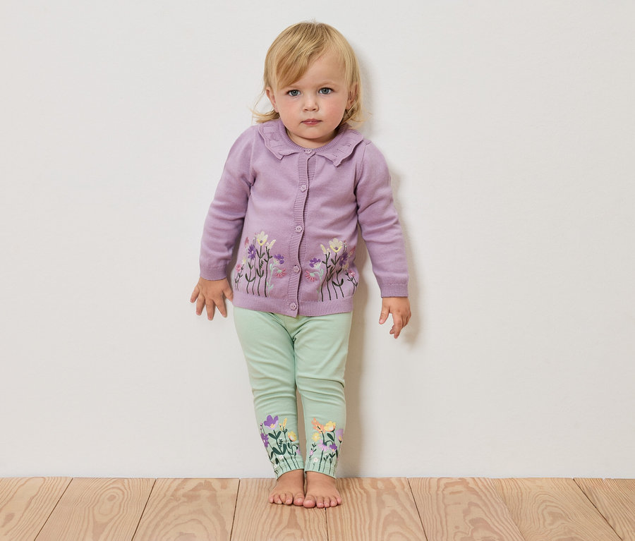 Egy szőke kislány mezítláb áll egy fehér fal előtt, lila, virágmintás kardigánt és zöld, virágos leggingset visel.