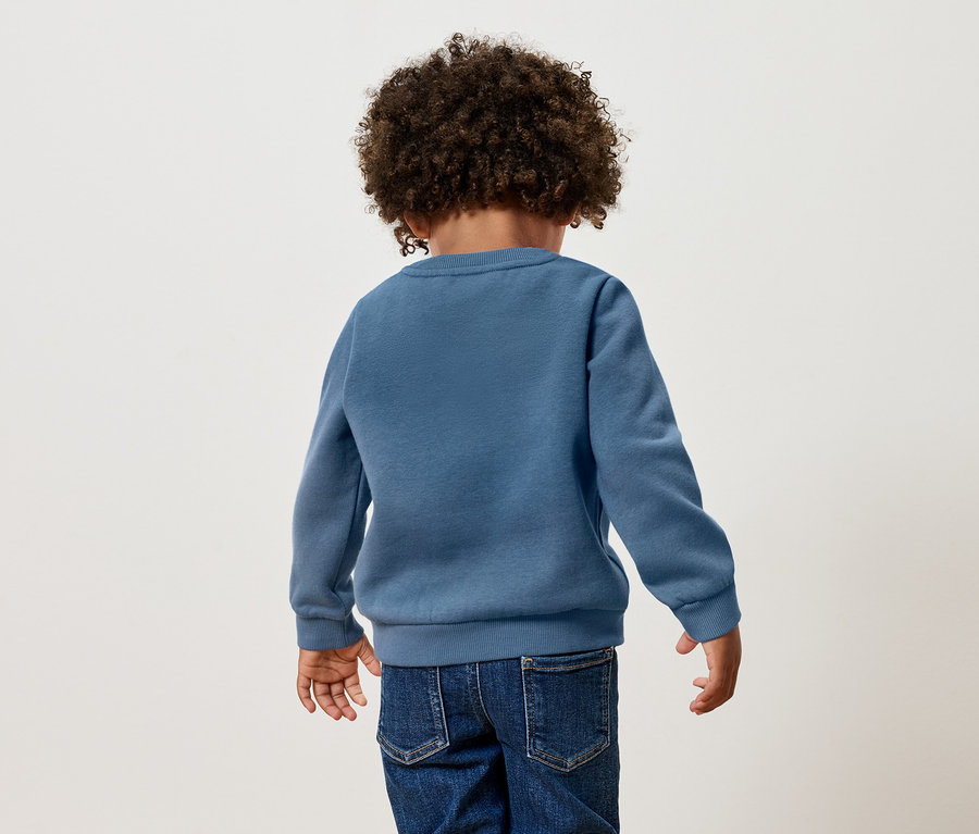 Hátulnézet egy göndör hajú gyermekről, aki kék pulóvert és farmert visel.