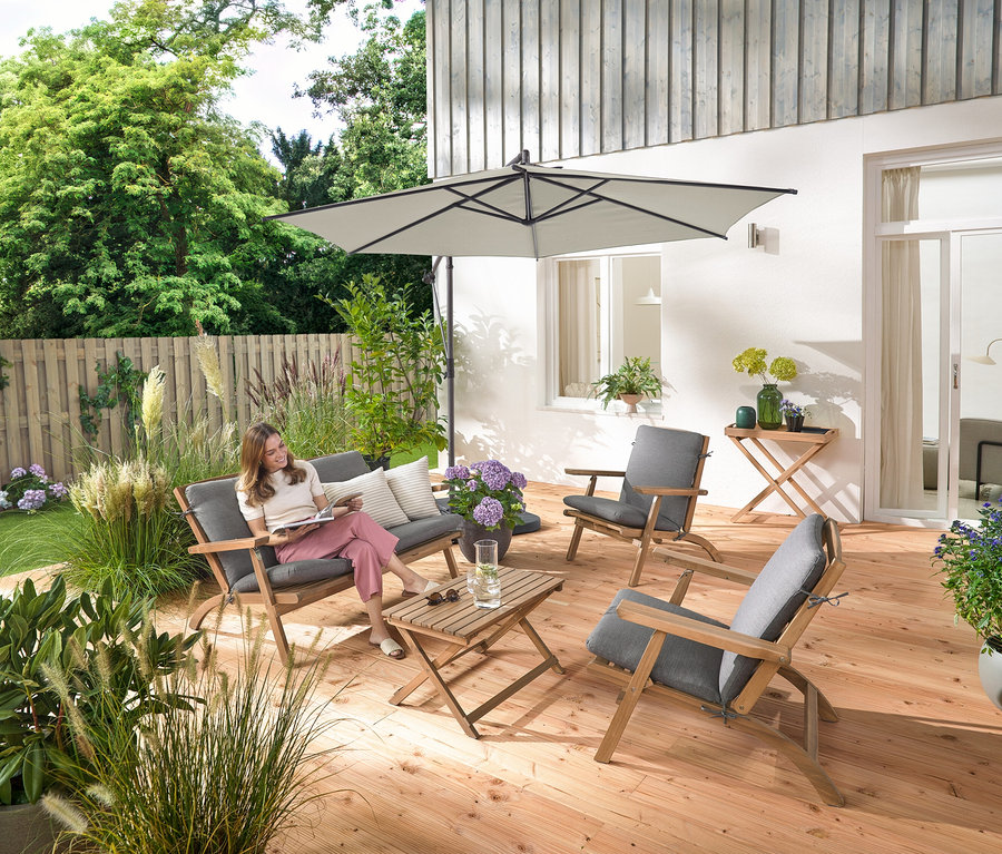 Egy nő ül egy összecsukható kerti társalgón prémium párnákkal, olvas, és körülötte egy kerti asztal és 2 vászon hatású kültéri párna található.
