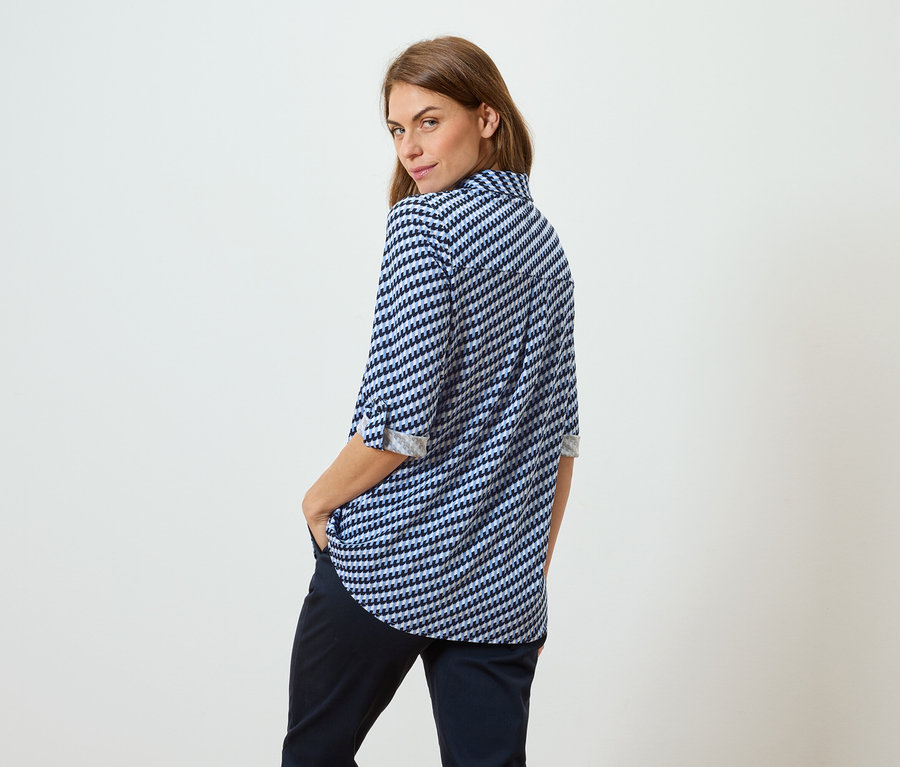 Barna hajú nő kék-fehér mintás blúzban és sötét nadrágban áll háttal a nézőnek, a válla fölött nézve.