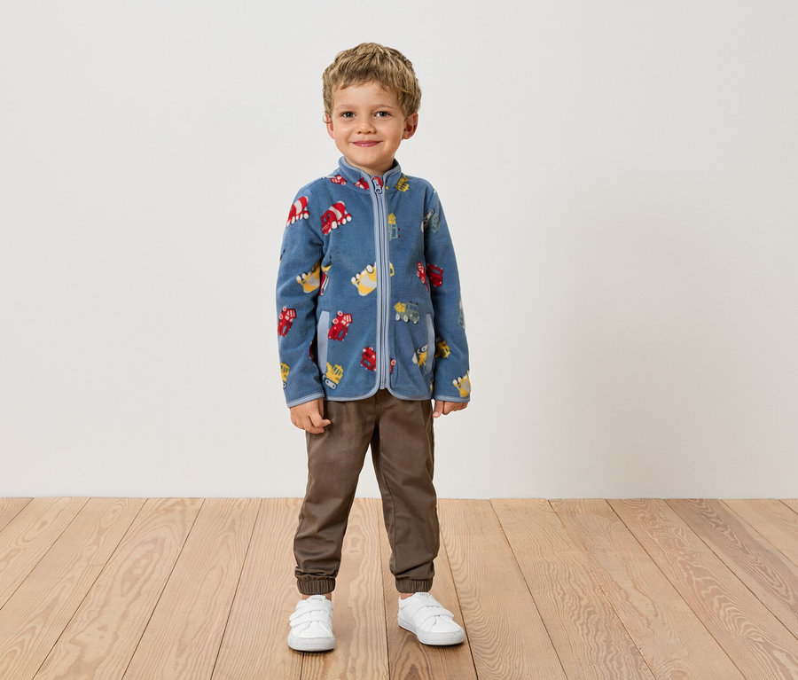 Egy fiú áll egy fal előtt, kék, járműmintás polárkabátban, barna nadrágban és fehér cipőben.