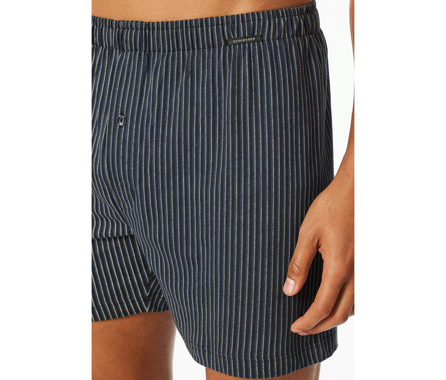 Közeli felvétel egy személyről, aki kék-fehér csíkos SCHIESSER jersey boxeralsót visel, gombos sliccel.