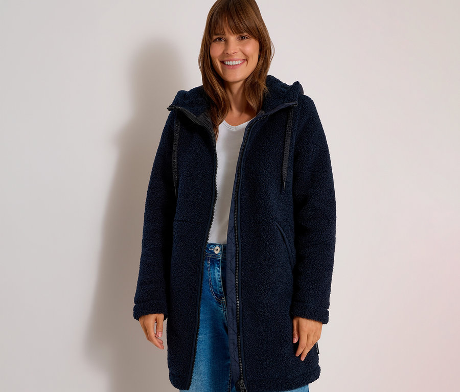 Mosolygó nő nyitott, sötétkék teddyfleece CECIL dzsekit, fehér pólót és farmert visel.