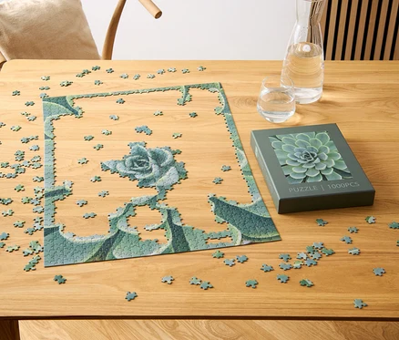 Egy részben összerakott, pozsgás növény mintájú puzzle egy fa asztalon van, egy pozsgás növény motívummal ellátott doboz, egy pohár és egy vízzel teli kancsó mellett.