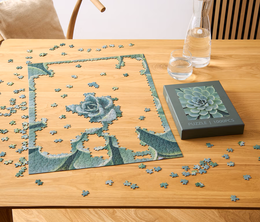 Egy részben összerakott, pozsgás növény mintájú puzzle egy fa asztalon van, egy pozsgás növény motívummal ellátott doboz, egy pohár és egy vízzel teli kancsó mellett.