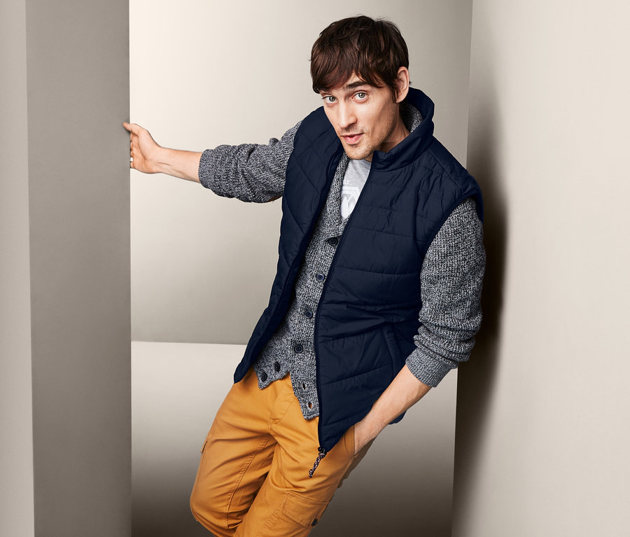 Férfi támaszkodik a falnak, steppelt mellényt, pulóvert és nadrágot visel.