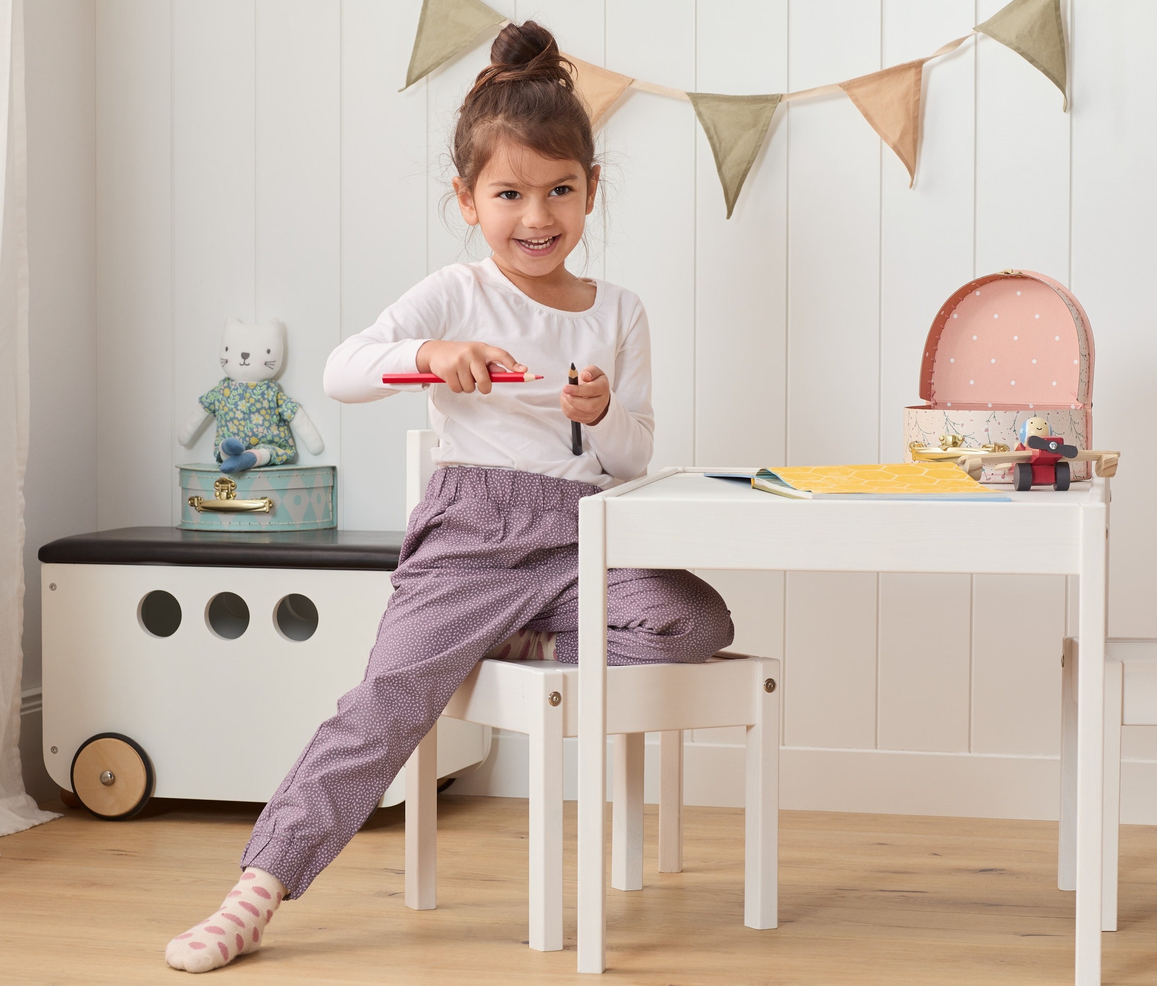 Kislány ül az asztalnál, ceruzákat tart. Mintás, lila kislány szabadidőnadrágot visel.