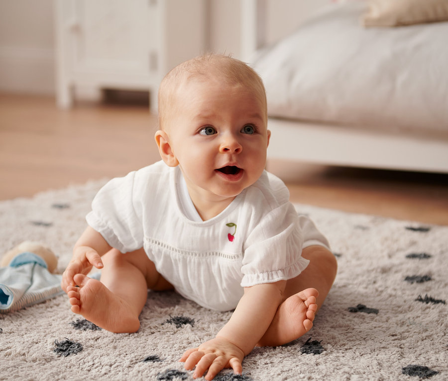 Egy baba ül a szőnyegen, és egy kislány rövid ujjú blúzt visel.