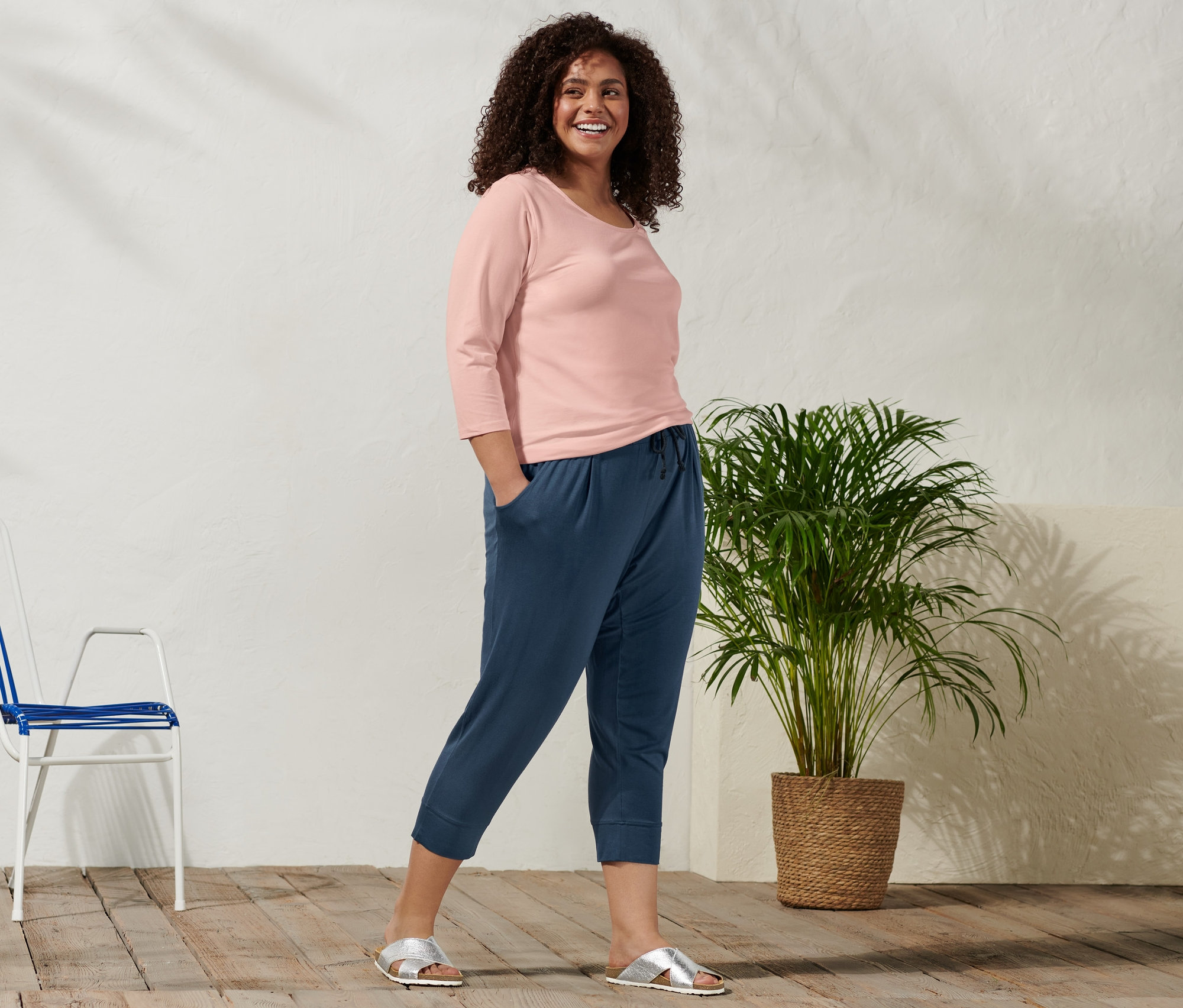 Mosolygó nő pózol rózsaszín, 3/4-es ujjú felsőben, sötétkék, 3/4-es dzsörzénadrágban és ezüst papucsban.