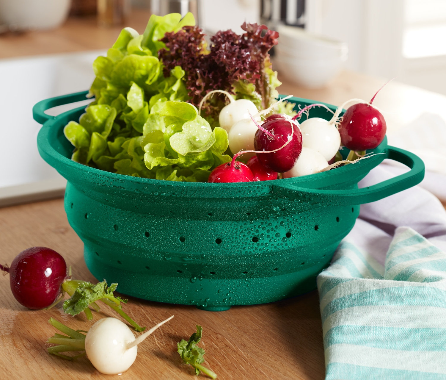 Egy zöld helytakarékos szilikon szűrő tele salátával és retekkel egy fa asztalon.