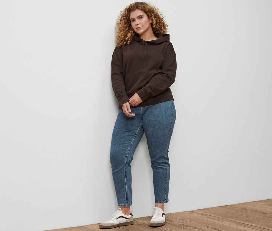Egy nő áll, rajta egyenes szárú »Fit Juna« farmernadrág, bőr sneaker és egy sötétbarna kapucnis szabadidőfelső.