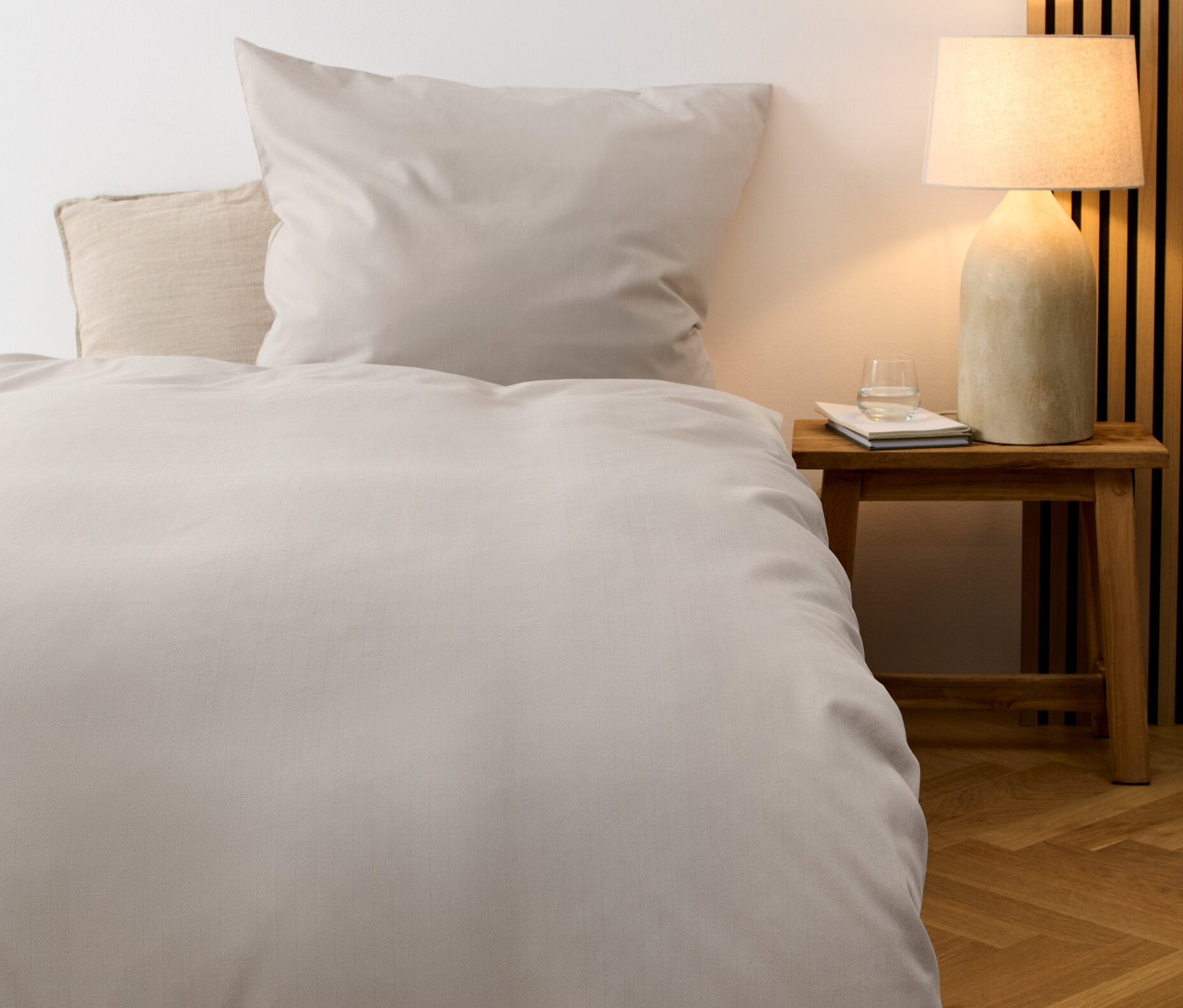 Egy világosszürke, normál méretű finom flanel ágynemű garnitúra van egy ágyon. Az ágy mellett egy éjjeliszekrény áll, rajta egy lámpa és egy pohár víz.