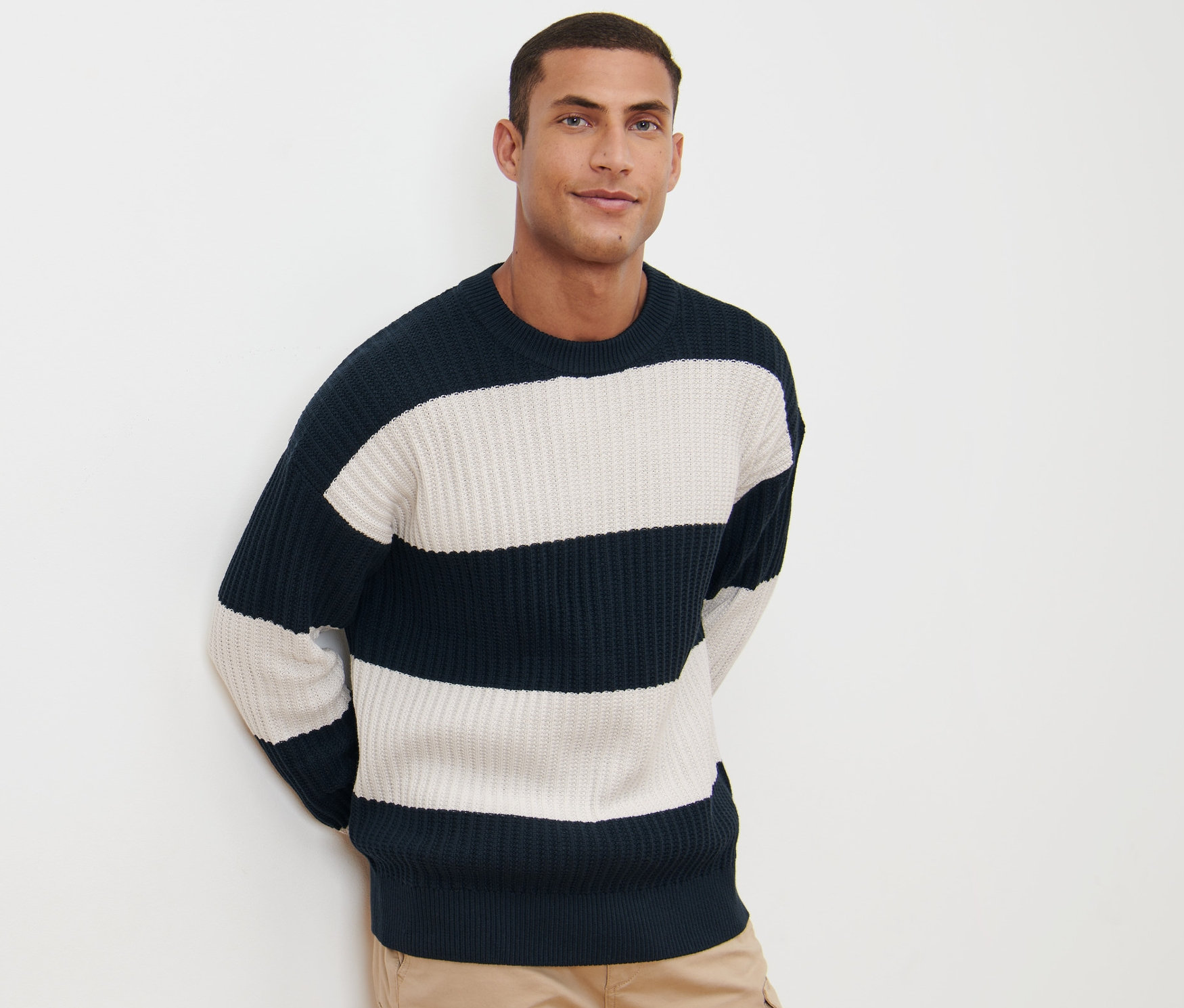 Férfi pózol fehér háttér előtt, tengerészkék-fehér csíkos, laza kötésű pulóvert és khaki nadrágot visel.