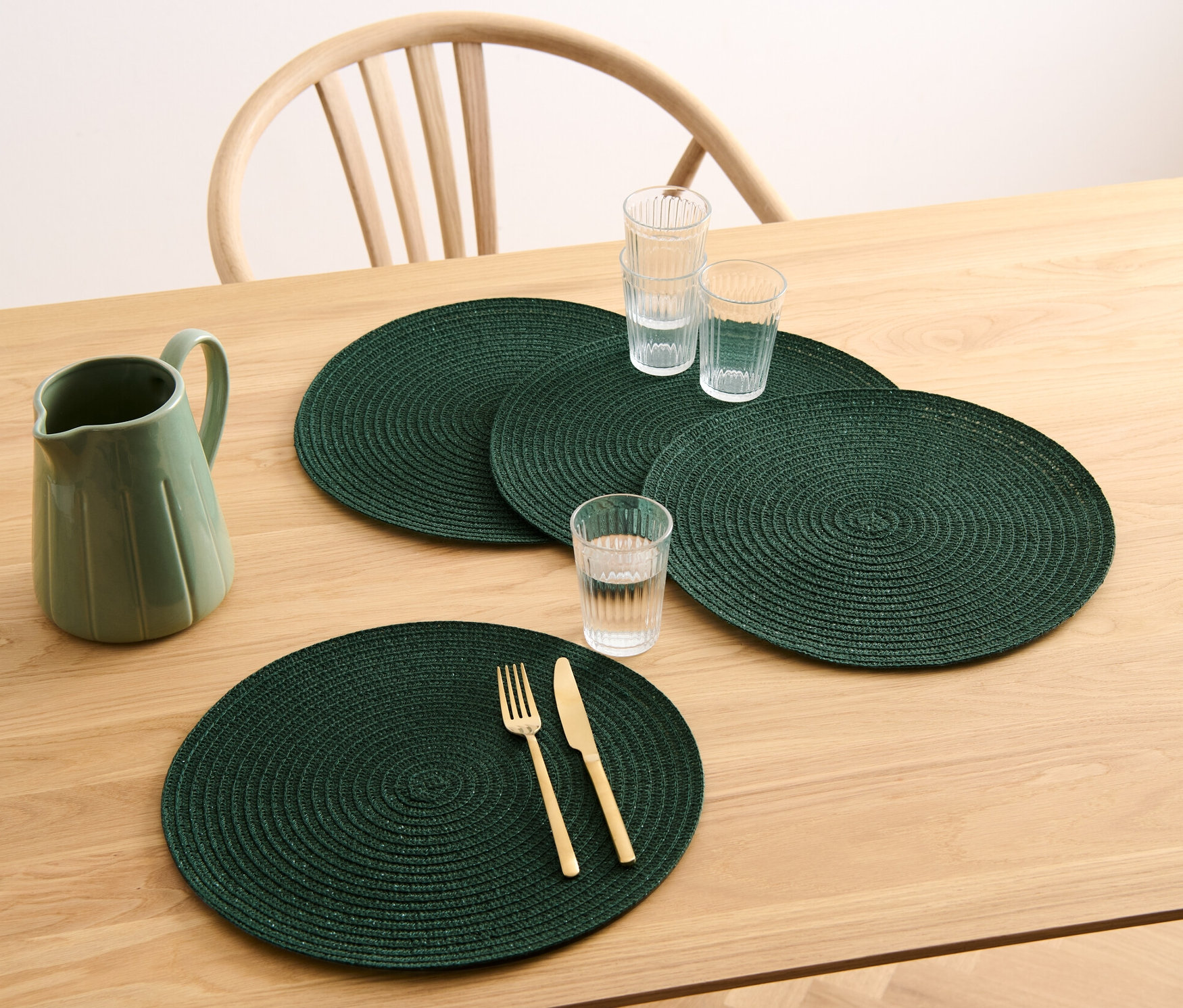 Egy fa asztalon négy zöld, kerek tányéralátét, egy kancsó és poharak vannak.