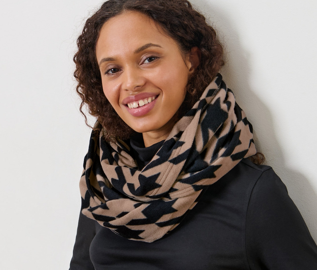 Barna, göndör hajú nő ül, tyúklábmintás női polár csősálat visel.