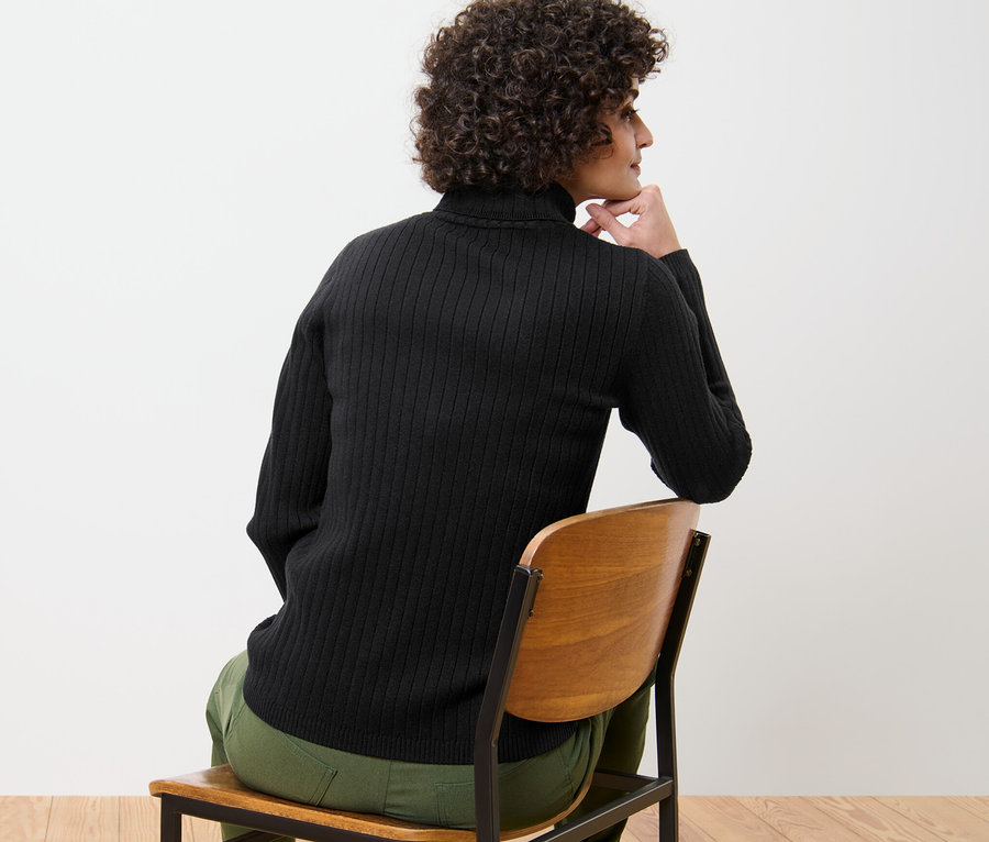 Egy nő ül egy széken, és fekete, garbónyakú, bordás kötésű pulóvert visel.