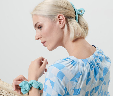 Egy szőke nő ül egy fotelben, a csuklóján egy hajgumi, a hajában pedig egy csat található. Frizurakészítő szett.